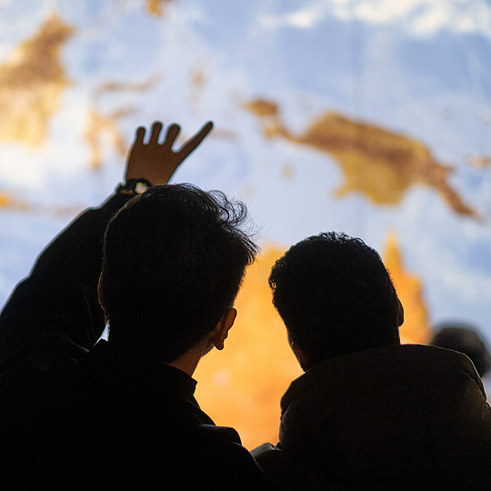 Zwei Menschen erscheinen als Silhouetten vor einer riesigen erleuchteten Erdkugel. Einer von Ihnen zeigt dabei auf die Erde. Zwei Menschen erscheinen als Silhouetten vor einer riesigen erleuchteten Erdkugel. Einer von Ihnen zeigt dabei auf die Erde.